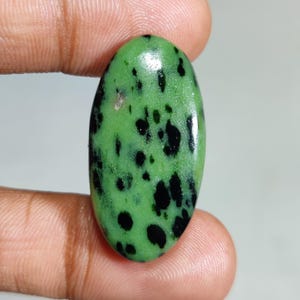 Puede incluir: Gema verde de forma ovalada con manchas negras. La piedra pulida se sostiene entre los dedos, mostrando su superficie lisa y su patrón natural. La gema es de un color verde vibrante.