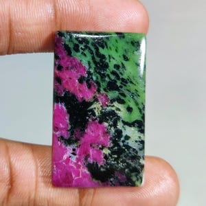 Peut inclure: Une pierre précieuse rectangulaire à surface polie. La pierre présente un mélange de couleurs vives, notamment du rose vif, du noir profond et du vert moucheté. La pierre est tenue entre les doigts, mettant en valeur sa taille et ses détails.