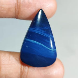 May include: A polished, teardrop-shaped gemstone with a deep blue base colour and swirling white and light blue patterns. The stone has a glossy finish and is held between fingers.
