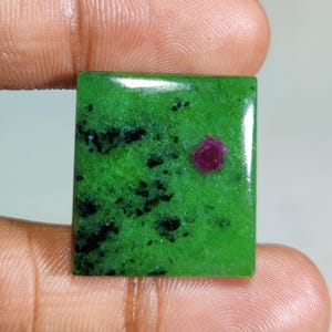 Puede incluir: Una gema de corte cuadrado con una base verde vibrante con motas negras y una inclusión roja oscura prominente. La superficie pulida refleja la luz, resaltando los patrones naturales y las variaciones de color. Ideal para la creación de joyas.