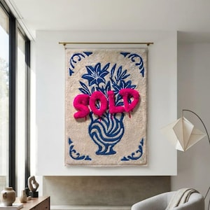 May include: A rectangular wall hanging featuring a beige tufted background with a blue line-art illustration of a vase filled with flowers. The word SOLD is written across the centre in raised, bright pink, puffy, textured lettering.