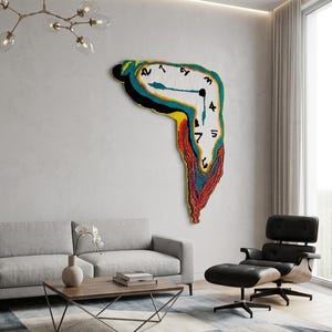 May include: A large, colorful wall hanging in the shape of a melting clock, reminiscent of a surrealist painting. The clock face is white with black numerals and hands, and the melting portion features vibrant hues of blue, yellow, and red. The artwork is displayed in a modern living room.