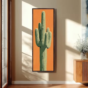 May include: A vertical rectangular wall art piece featuring a saguaro cactus illustration centered on a solid orange background. The cactus is rendered in shades of light and dark green with visible vertical ribs and multiple upward-reaching arms.