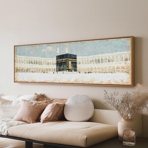 May include: A panoramic wall art print featuring a painterly depiction of the Kaaba in Mecca. The artwork showcases the black and gold structure centered against a pale blue sky and arched colonnades, all enclosed within a light wood frame.