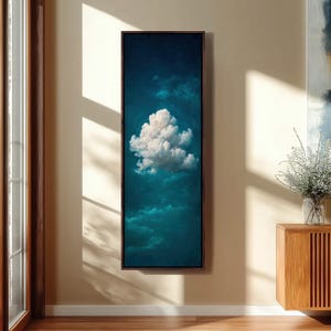 Puede incluir: Una obra de arte vertical enmarcada que representa un paisaje de nubes. La obra presenta una gran nube blanca y esponjosa sobre un fondo de tonos azul verdoso y azul oscuro. El marco es de color marrón oscuro y la obra se exhibe en una pared beige.