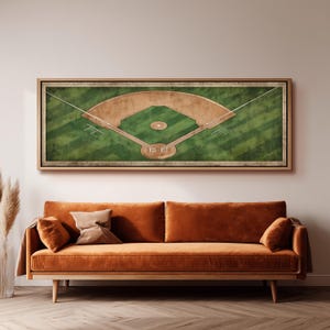 May include: A framed baseball field art print. The artwork depicts a detailed overhead view of a baseball field, with green grass, brown dirt, and white lines. The frame is a light brown color. The artwork is displayed above an orange sofa.