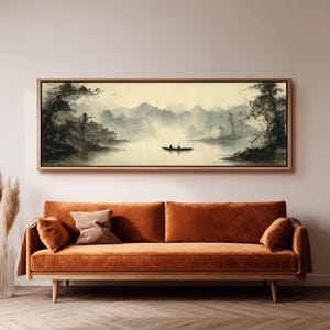Puede incluir: Una pintura de paisaje panorámico en un marco de madera clara que muestra una escena de lago brumoso con montañas, árboles, un pequeño edificio en la orilla y un bote con dos figuras en el agua, sobre un sofá largo de terciopelo color naranja.