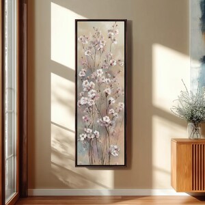 以下が含まれることがあります： 垂直に額装されたキャンバスアート作品で、花のモチーフが描かれています。 このアートワークは、ベージュ、ピンク、ブルーの柔らかく落ち着いた背景に、細い茎に繊細な白い花とピンクのアクセントを特徴としています。 フレームは濃い茶色です。
