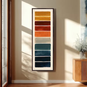 May include: A vertical rectangular wall art piece featuring a series of nine stacked horizontal color blocks in shades of yellow, orange, red, gray, and blue, all enclosed within a thin dark brown frame against a light wall.