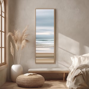 Puede incluir: Una obra de arte enmarcada, rectangular y alargada, que representa una escena costera borrosa. La obra presenta bandas horizontales de color, incluyendo tonos de azul, beige y blanco, evocando el mar y el cielo. El marco es de madera clara, en una habitación de tonos neutros.