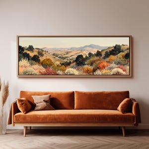 May include: A framed landscape painting shows a hillside with trees and bushes in green, orange, and red hues. The artwork hangs above a rust-coloured velvet sofa with wooden legs, creating a warm atmosphere. The painting's frame is made of wood.