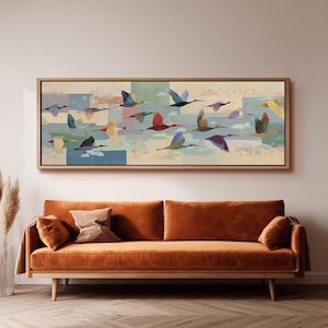 Puede incluir: Una obra de arte en lienzo enmarcada rectangular que representa una bandada de pájaros en vuelo. Las aves están representadas en varios colores, incluyendo azul, rojo y amarillo, contra un fondo de nubes y bloques de colores pastel. La obra de arte se exhibe sobre un sofá naranja.