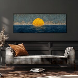Op de afbeelding: Een ingelijst kunstwerk dat een zonsondergang boven de oceaan afbeeldt. De zon is een heldergele bol, die reflecteert op het blauwe water. Daaronder een zwart-wit patroonbank met een bruin kussen.