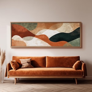 May include: A rectangular wall art piece in a light wood frame featuring an abstract landscape with flowing, layered shapes in shades of terracotta, sage green, cream, and dark forest green, displayed above a long, solid burnt orange velvet sofa.