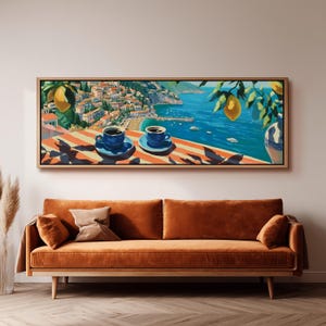May include: A framed art print showing a coastal scene with two blue coffee cups on a striped table. The artwork features a vibrant blue sea, a town on a hillside, and lemon branches. The frame is a light wood colour.