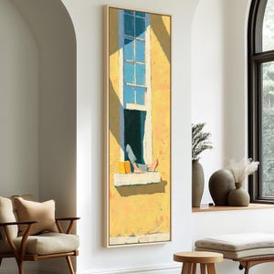 May include: A vertical framed wall art print depicting a yellow building facade with a tall window. A person wearing blue jeans sits on the window ledge, reading a yellow book with bare feet resting on the white sill under bright sunlight.