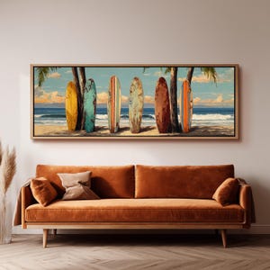 May include: A framed canvas print showing six surfboards on a sandy beach, leaning against palm trees. The surfboards are in various colours, including yellow, teal, and brown. The ocean and sky are in the background, with a burnt orange sofa in the foreground.