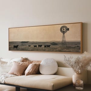 Könnte beinhalten: Ein panoramisches, rechteckiges Wandbild in einem hellen Holzrahmen mit einer Landschaft in Sepiatönen. Die Szene zeigt ein weites Grasfeld mit fünf dunklen Silhouetten von weidenden Rindern und einer hohen, metallenen Windmühle.