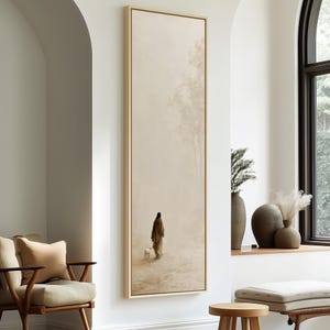 Peut inclure: Une impression sur toile verticale et rectangulaire dans un cadre en bois clair, représentant une peinture beige monochrome et atténuée d'une silhouette solitaire en longue robe marchant dans un paysage vaste et brumeux avec un petit animal blanc.
