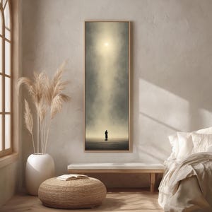 Puede incluir: Una impresión artística vertical enmarcada representa una figura solitaria en un paisaje brumoso, iluminada por una luz brillante. La obra de arte está en una paleta de colores neutros, con un marco de madera.