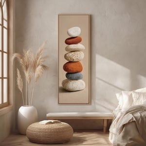 May include: A framed vertical artwork featuring a stack of smooth, colorful stones. The stones are in shades of white, orange, gray, and beige. The artwork is displayed in a neutral-toned room with a window, a bench, and a round ottoman.