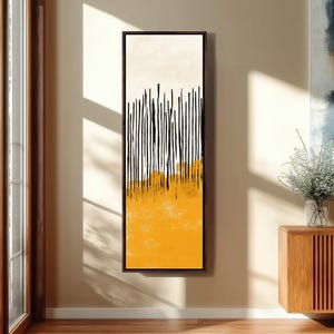 Può includere: Stampa artistica astratta in una cornice marrone scuro. L'opera presenta linee nere verticali su uno sfondo giallo e crema. Il pezzo è montato su una parete beige, con un mobile in legno e una finestra visibili sullo sfondo.