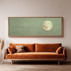 Könnte beinhalten: Ein gerahmtes Kunstwerk, das einen Vollmond und einen Vogelschwarm im Flug vor einem gedämpften grünen Himmel zeigt. Das Kunstwerk hängt über einem rostorangen Samtsofa mit Holzbeinen und schafft eine warme und einladende Ästhetik.