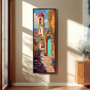 May include: A vertical framed wall art print depicting a sunlit Mediterranean street scene with a tall bell tower, a bright turquoise arched door, and vibrant orange stucco walls. The path is paved with colorful tiles and lined with potted plants.