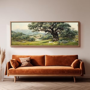 May include: A panoramic landscape painting in a light wood frame featuring a large, gnarled oak tree in a grassy meadow with rolling hills and distant mountains under a soft sky. The artwork is displayed above a long, burnt orange velvet sofa.
