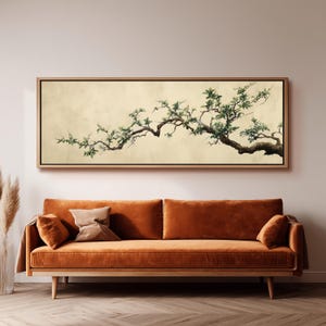 Puede incluir: Una impresión artística enmarcada que presenta una ilustración detallada de una rama de árbol con hojas verdes y un tronco marrón. La obra de arte está sobre un fondo crema y enmarcada en madera clara. La impresión cuelga sobre un sofá naranja.