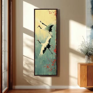 Puede incluir: Una pieza de arte mural rectangular vertical que muestra dos grullas blancas en vuelo con puntas de alas negras y coronas rojas. El fondo transita de un amarillo pálido arriba a un verde azulado apagado abajo, con ramas de flores rojas.