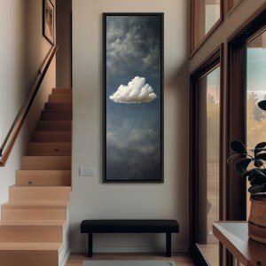 Könnte beinhalten: Ein gerahmter, vertikaler Kunstdruck mit einer einzelnen, flauschigen weißen Wolke vor dem Hintergrund eines düsteren, dunkelblauen Himmels. Das Kunstwerk wird an einer Wand neben einer Holztreppe und einer schwarzen Bank ausgestellt.