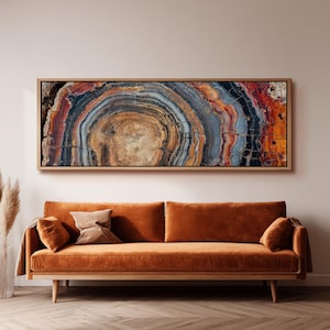 Puede incluir: Una impresión enmarcada de arte mural que presenta una sección transversal de un tronco de árbol, mostrando anillos concéntricos en tonos marrón, naranja, azul y rojo. La obra de arte se exhibe sobre un sofá de terciopelo naranja quemado con patas de madera.