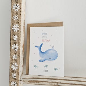 Könnte beinhalten: Geburtstagskarte mit einer Aquarellillustration eines blauen Wals und drei kleinen Fischen. Auf der Karte steht "Happy Happy Birthday" mit dem Namen "Liam" darunter. Die Karte befindet sich vor einem neutralen Hintergrund.