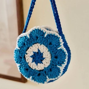 Puede incluir: Bolso de ganchillo hecho a mano en tonos azules y blancos, con un diseño floral en la parte delantera. El bolso tiene forma redonda y una correa azul para llevarlo. El trabajo de ganchillo es detallado.
