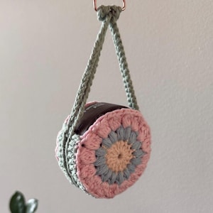 Può includere: Un portavaso all'uncinetto fatto a mano di forma circolare, sospeso da un cordoncino verde intrecciato e un anello color rame. Il supporto presenta un motivo floreale rosa, grigio e beige, perfetto per piccole piante o oggetti decorativi.