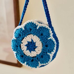 May include: A handmade crochet bag in shades of blue and white. The bag features a floral design on the front and a matching blue strap. The bag is round and appears to be small, perfect for carrying essentials.