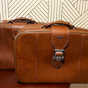 May include: Two rectangular brown suitcases with a smooth, leather-like finish. Each suitcase features a top handle, a central vertical strap with a silver-toned metal buckle, and a small rectangular logo plate labeled Meridian on the front.