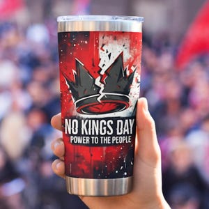 Peut inclure: Un gobelet en acier inoxydable avec un design rouge et blanc, orné d'un graphisme de couronne brisée et du texte "NO KINGS DAY POWER TO THE PEOPLE". Le gobelet est tenu devant une foule floue.