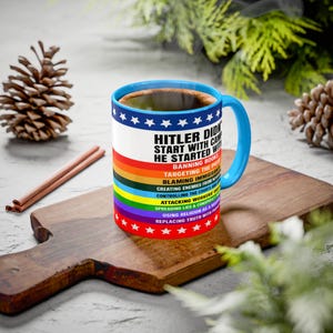 May include: A ceramic coffee mug with a blue handle and rim, filled with coffee. The mug features text in white and a rainbow design. The text reads "HITLER DID START WITH...HE STARTED WITH...BANNING BOOKS..." and more. The mug sits on a wooden cutting board.