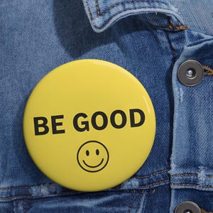 Pode incluir: Um botão amarelo brilhante com as palavras "BE GOOD" em letras pretas e uma carinha sorridente. O botão está preso a uma jaqueta jeans azul, criando uma mensagem positiva e alegre.