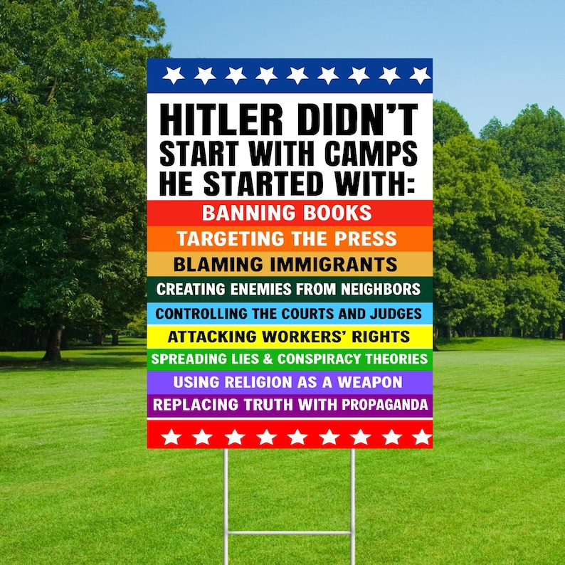 May include: A rectangular yard sign with a white background and text that reads "HITLER DIDN'T START WITH CAMPS HE STARTED WITH:" followed by a list of actions. The sign has a blue top with white stars and a red bottom with white stars.