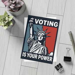 May include: A greeting card with the Statue of Liberty illustration, with the words "VOTING IS YOUR POWER" in white text. The background is divided into red and blue sections. The card is on a white surface with office supplies and potted succulents.
