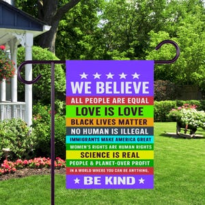 May include: A garden flag with a purple background and white text that reads "WE BELIEVE" and other statements about equality and kindness. The flag is displayed on a dark metal stand in a green yard.