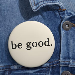 May include: A round, off-white button with the phrase "be good." in black, sans-serif font. The button is pinned to a blue denim jacket, with visible stitching and a button closure.