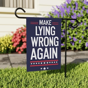 May include: A navy blue garden flag with the words "MAKE LYING WRONG AGAIN" in white. The flag has red stripes and stars at the top and bottom. It is displayed on a black metal stand in a garden setting.