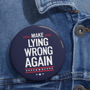 May include: A round, navy blue button with the text "MAKE LYING WRONG AGAIN" in white. The button has red stripes and stars. The button is pinned to a blue denim jacket.