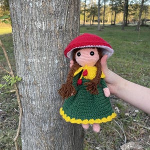 May include: A handmade crochet doll with a red mushroom cap, a yellow top, and a green dress with yellow trim. The doll has brown braids and pink feet. The background shows a tree and a grassy area.