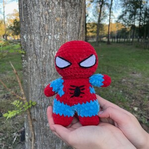 May include: A handmade plush Spider-Man doll. The doll features a red and blue color scheme, with white eye details and a black spider emblem on the chest. The doll is held in front of a tree.