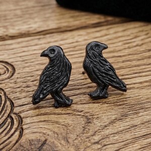May include: Two black raven-shaped stud earrings. The earrings are displayed on a wooden surface with a decorative carved design. A black jewelry box is in the background. The earrings are a dark, matte black.